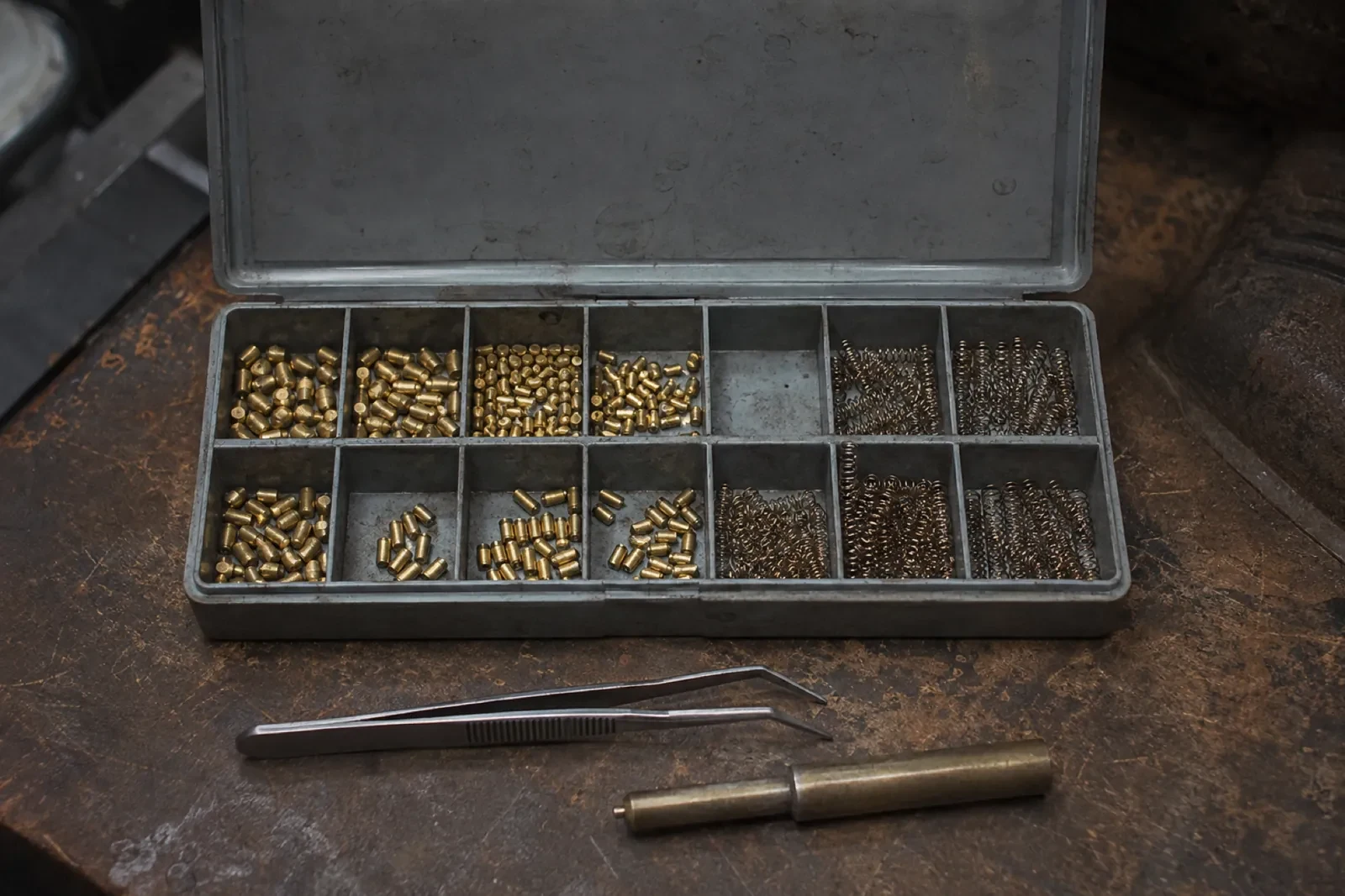 Open metal case with brass pins and coiled springs sorted in small sections, with tweezers and a brass tool in front. — Eringate
